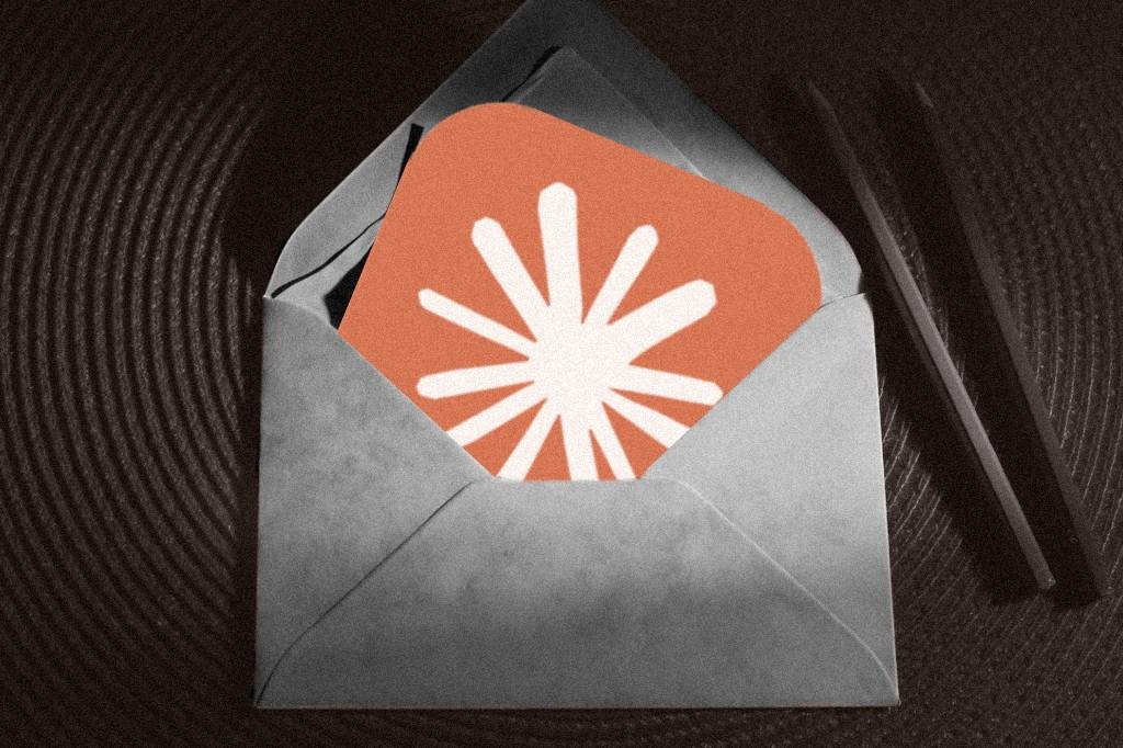 Envelope cinza aberto com um cartão laranja dentro, exibindo um símbolo branco de estrela ou floco de neve com oito pontas. À direita, dois lápis escuros sobre uma superfície texturizada em tons de marrom. A imagem tem um filtro granulado e escuro