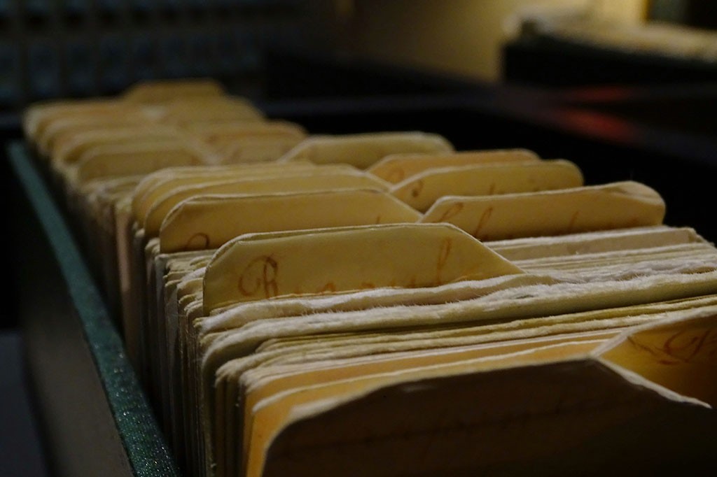 Fichas de catálogo amareladas, com abas de identificação e letras manuscritas, organizadas em uma gaveta escura de arquivo, vistas em perspectiva diagonal