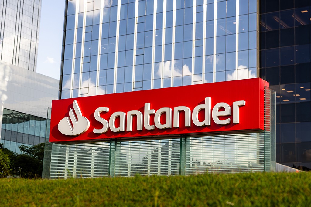 Letreiro vermelho com o nome Santander em branco e o logotipo do banco, em frente a um prédio moderno de vidro espelhado, com gramado verde em primeiro plano